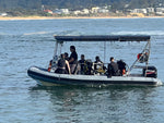 A white and blue rigid airboat with a black canopy is floating on the water, carrying several people equipped with scuba gear. A sandy beach and coastal buildings are visible in the background.